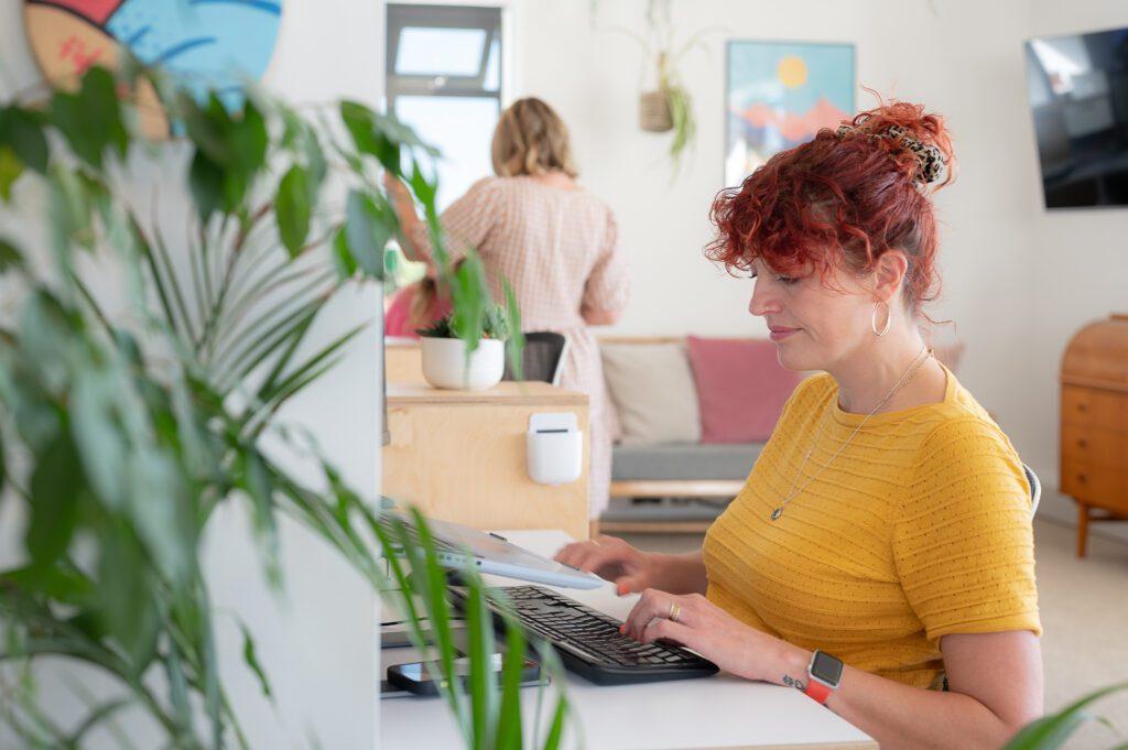 SEO Content Creation That Connects Users and Search Engines 2 Red haired lady creates SEO content sat at a white desk with plants around her.