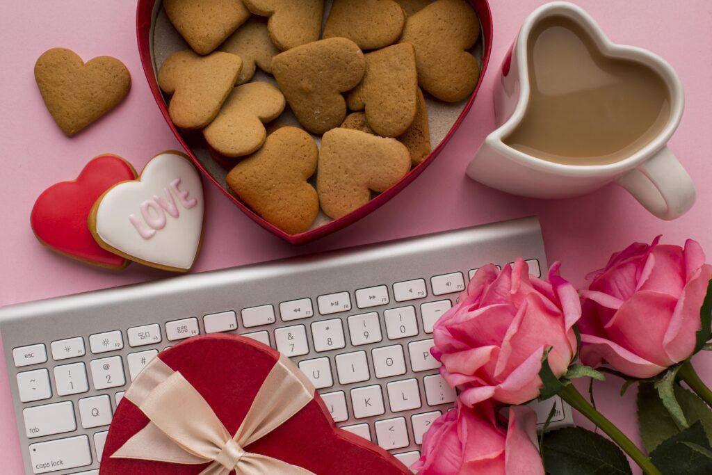 A white computer keyboard covered in roses and heart shaped biscuits and tea for valentines day.