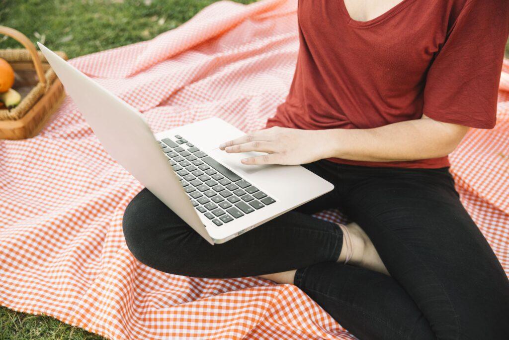 A close up of a laptop on a womans lap, sat crossed legged on a red gingham picnic blanket in the summer.