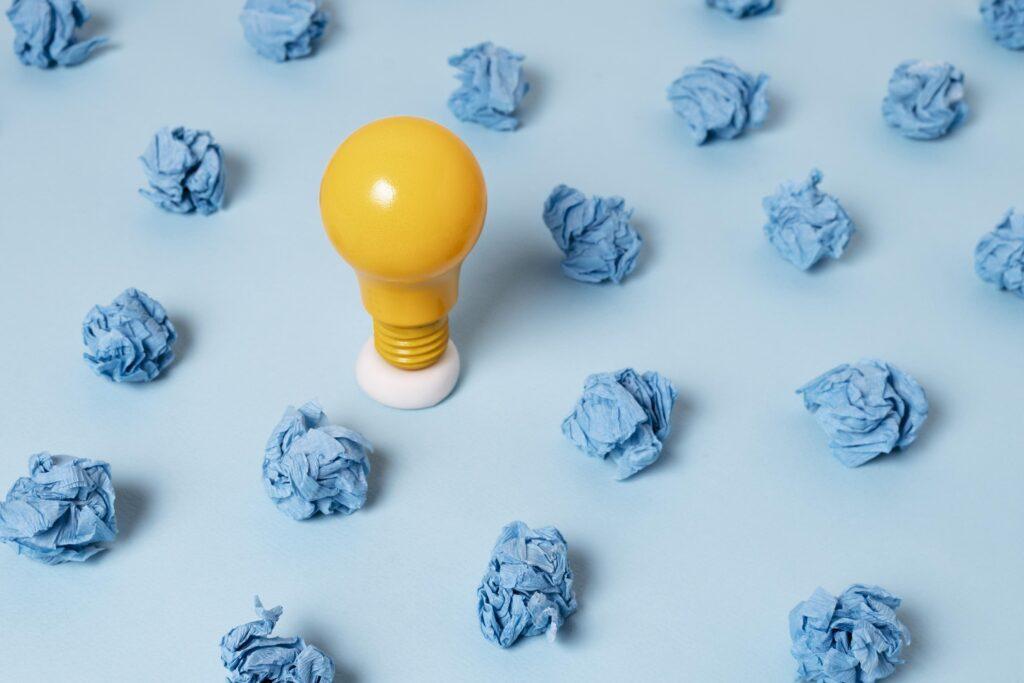 Yellow light bulb on a blue paper background surrounded by crumpled balls of blue paper