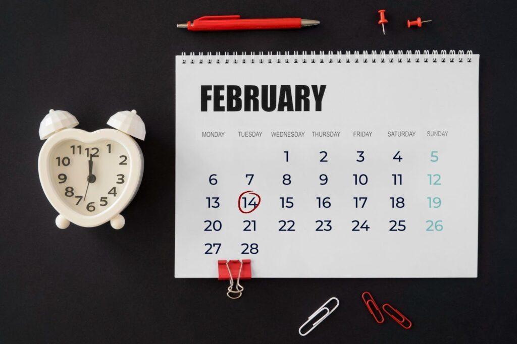 A calendar set on the month of February, circling the 14th (Valentines day). A clock and paper clips surround the calendar.