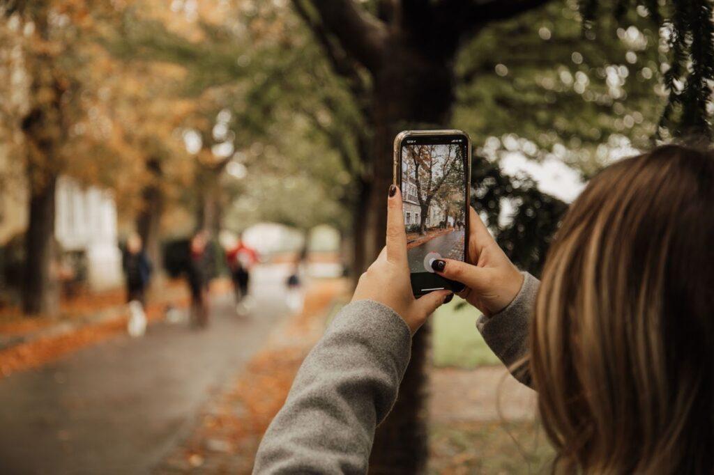 A girl holds up her phone camera to take a photo of an autumnal road with orange leaves on the ground for content creation.