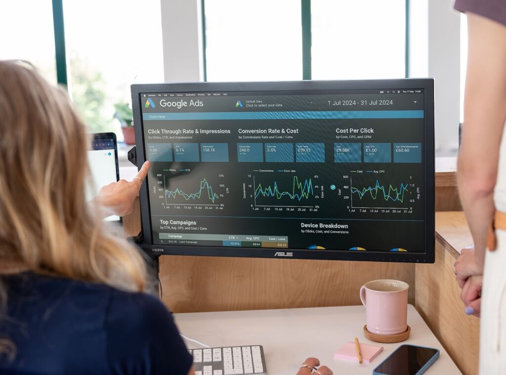 Over the shoulder shot of a blonde girl pointing at PPC results from Google Ads. There is a pink mug on the table and pink sticky notes.