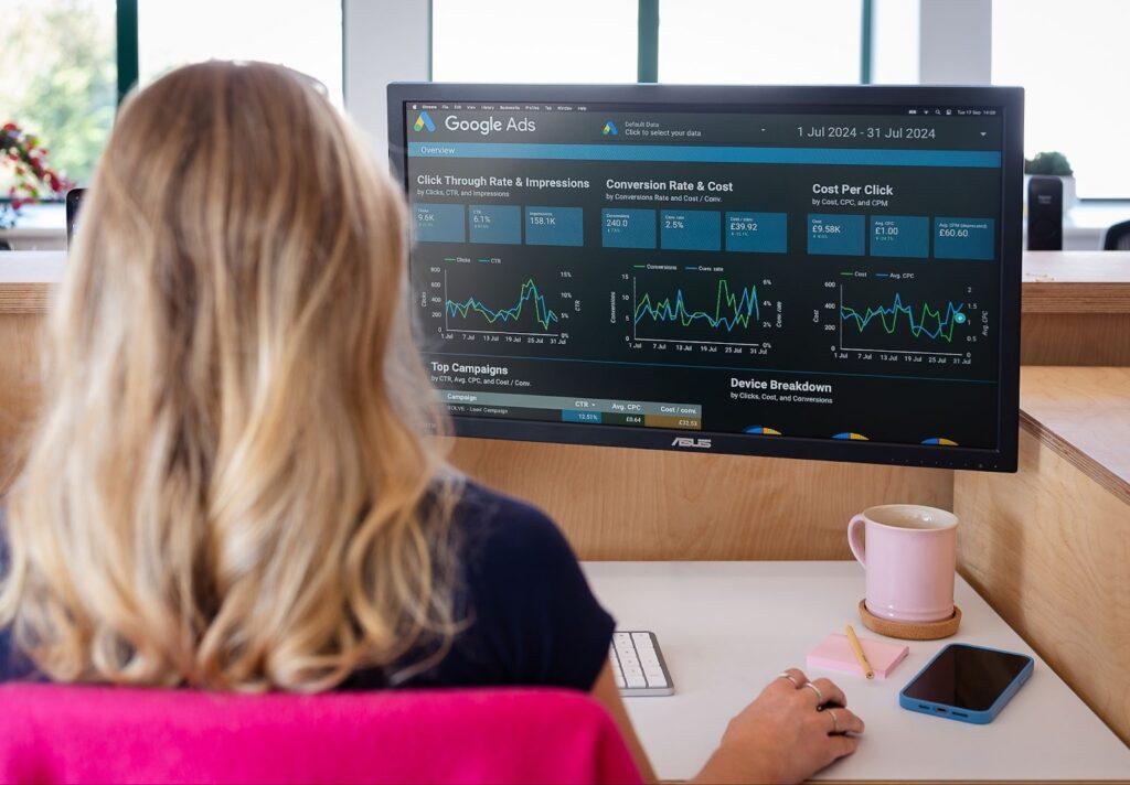 11 Common PPC Mistakes to Avoid in Your Next Campaign 3 Blonde girl sits at her desk looking at a computer screen showing pay per click results on google ads, with a pink mug on the desk.