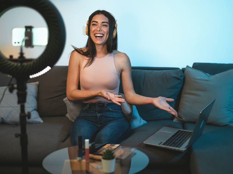 A female influencer sitting on her sofa with a laptop to her side, wearing headphones. A mobile phone is on a stand filming her whilst she speaks.