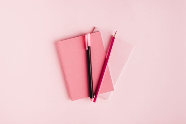 Pink paper background with two pink planners laying on one another, with a pink pen and pencil on top representing content planning tips.