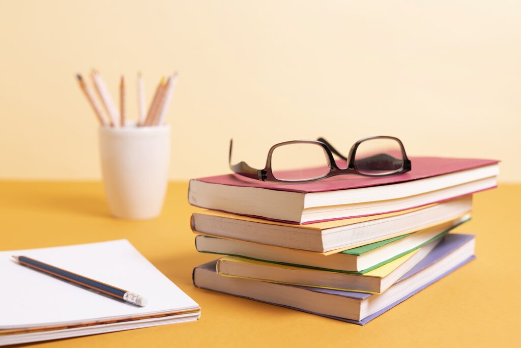 Stacks of books with glasses placed on top, sat on a yellow table with pens and pencils in the background.