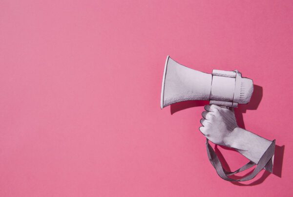 A pink background with a paper drawing of a hand holding a megaphone up to represent your social media brand voice.