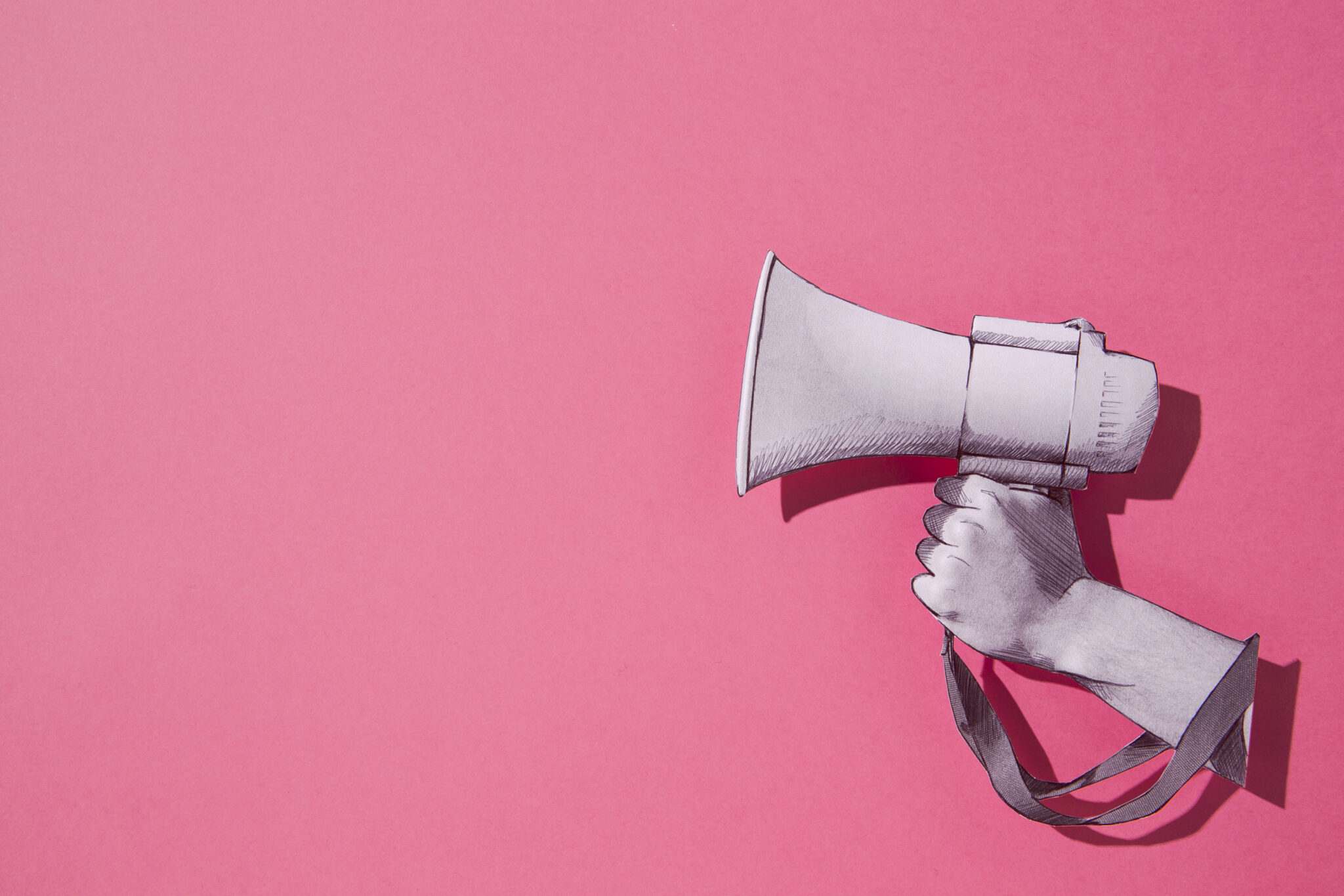 Why Your Social Media Brand Voice Matters More Than You Think 1 A pink background with a paper drawing of a hand holding a megaphone up to represent your social media brand voice.